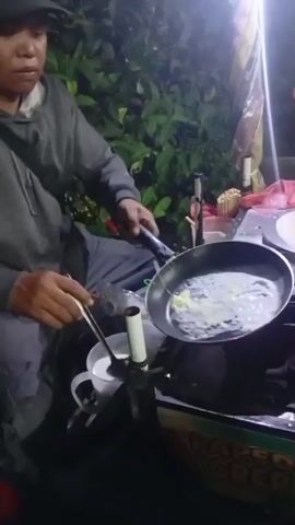 A person wearing a gray hooded sweatshirt and a dark baseball cap is cooking in a pan on a portable stove. The person is holding a utensil in their right hand and is manipulating something in the pan with it. The pan contains a liquid and appears to be heating up. There is a white mug with liquid next to the stove, and an umbrella is also visible. The background is dark and shows some greenery, suggesting an outdoor setting at night. The person seems to be talking or singing, with their mouth open and teeth visible.