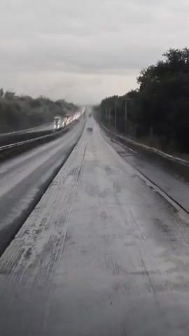 The video is a first-person perspective shot from a vehicle traveling on a highway. The road appears wet, possibly from rain, and there is visible traffic in the distance. The sky is overcast and gray. The focus is on the road ahead, with trees lining the sides of the highway. There are tire tracks visible on the wet surface of the road.