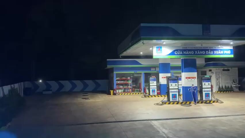 The video shows a gas station at night. There are several gas pumps under a brightly lit canopy. A person in a blue uniform is standing near one of the pumps. A car is parked to the right of the gas station. The background is dark, suggesting it is nighttime. The gas station appears to be open for business.