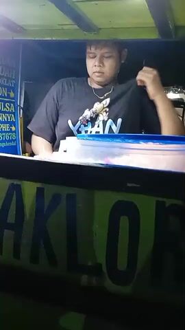 A person is standing inside a food cart, preparing food. They are wearing a black t-shirt with a cartoon print and a silver necklace. The cart has a green and yellow sign that reads "MAKLOR". Behind them is a sign with text written in Indonesian, which seems to list the items being sold. They are reaching for a yellow container on the counter. The environment is dimly lit.
