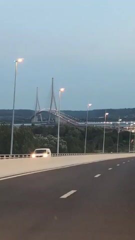 The video shows a view from a moving vehicle on a highway. The highway has multiple lanes and is lined with streetlights. In the distance, a large cable-stayed bridge can be seen. The sky is overcast with a pale blue color, suggesting either dawn or dusk. Trees and a body of water are visible below the bridge. The vehicle is traveling towards the bridge.