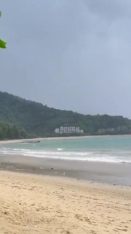 A sandy beach is in the foreground with small waves rolling onto the shore. In the middle ground, the turquoise ocean stretches to the horizon. Lush green hills covered in trees form the background, with a few white buildings visible on the hillside. The sky is overcast and appears to be clearing up. A few boats are visible in the water. The video appears to be shot on a cloudy day with signs of recent rain.