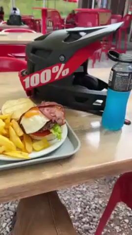 The video shows a lunch break in a mountain resort. A table in an outdoor seating area has a grey tray containing a plate with a burger topped with a fried egg and bacon, a side of French fries, cutlery, and a ketchup packet. A mountain bike helmet sits next to the tray. The camera pans out to show other red tables and chairs, people walking around with bicycles, and a view of a grassy area with mountains in the background. There's also a ski lift visible.