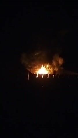 The video shows a fire burning in a grill at night. Flames and smoke are visible, illuminating the dark surroundings.