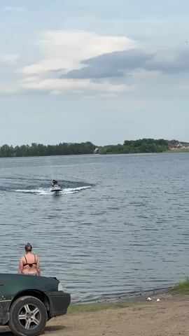 A person rides a jet ski across a body of water, leaving a wake behind them. In the foreground, a person sits on the hood of a green vehicle facing away from the camera. Further along the shore, two people stand near the water's edge, and a child sits on a yellow towel. The scene takes place on a cloudy day with a tree line and buildings visible in the distance across the water.