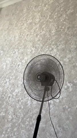 A dark-colored oscillating fan stands against a textured grey wall in this video. The fan is on, and its circular grill is visible. The fan oscillates from left to right.