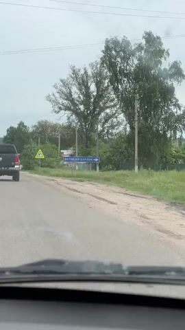 The video shows a car driving on a road. The car passes a road sign that reads "ВЕЛИКИЙ БУРЛУК" with an arrow pointing to the right. There are trees and power lines visible in the background. Another car is ahead on the same road. The weather appears overcast.