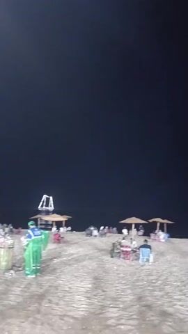A wide shot shows a beach at night with a dark sky. In the foreground, a person in a green uniform is cleaning a trash can. They are wearing gloves and a face mask and appear to be collecting litter. Several beach umbrellas are set up in the background, and a few people are seated under them. Further back, there is a structure with lights, possibly a decorative archway. The sand is visible, and there are some objects scattered on it. The overall impression is of a beach being cleaned after a day of activity.