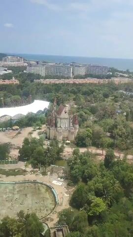 The video presents a high-angle view of a run-down area near a coastline. In the foreground, a large, empty, circular pool with discolored concrete and visible drain holes takes up a significant portion of the frame. Next to the pool, dense greenery with various trees and vegetation surrounds the area. A prominent feature is a dilapidated castle-like structure with visible signs of age and disrepair, including faded colors and weathered surfaces. A large white tent-like structure is also visible in the vicinity.  Further in the background, numerous buildings, possibly hotels or resorts, line the coastline. Beyond the buildings, the ocean stretches to the horizon under a bright, partly cloudy sky. No people are visible in the scene.