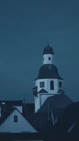 The video shows a time-lapse of a clock tower against a dark blue sky. The clock faces are visible and appear to be digital or analog with hands. The sky transitions from a lighter shade of blue to a darker shade, indicating the passage of time from dusk into night. Buildings with pitched roofs are visible in the foreground, partially obscuring the base of the clock tower. There are no people or any other discernible activity in the video.