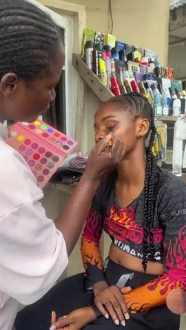 In this video, a makeup artist is applying eyeshadow to a young woman's right eye. The makeup artist is holding an eyeshadow palette with many different colors and a brush, carefully applying the makeup. The woman has her eyes closed and is wearing a black cropped shirt with a flame design. She has her hair braided into two long pigtails. The background shows a stall filled with various cosmetic products, indicating an outdoor market or shop setting. The lighting is natural, suggesting the video was filmed during the daytime.