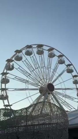 A Ferris wheel stands against a pale blue sky.  The wheel is white with glass enclosed passenger cars.  The center of the wheel features a red circle.  The bottom of the frame is partially obscured by what appears to be a wire mesh structure with small lights.