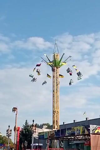 A tall, yellow amusement park ride with a green top is in motion against a blue sky with scattered clouds. Several gondolas are suspended from the arms of the ride, each carrying passengers who are sitting and looking in various directions. Some of the gondolas are adorned with flags, including what appear to be Ukrainian flags. In the background, other amusement park structures, including a red ride, are visible, along with trees and a building with signage. The ride appears to be in operation, with the gondolas swinging outwards and upwards as the central mechanism rotates. The lighting suggests it is daytime.