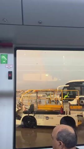 The video shows a view from inside an airport bus. Through the bus window, a rainbow is visible in the distance, above airport tarmac. On the tarmac, an airport tug vehicle is parked, with its yellow safety bars and equipment visible. A man is seated at the front of the bus, partially visible in the lower right corner of the frame. The weather appears overcast and wet. The bus window has condensation and raindrops on it, creating a blurred effect. The camera pans upward, focusing on the rainbow and the aircraft shadow.