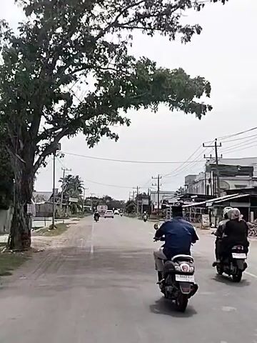 The video shows a street scene with two people riding motorcycles. The first motorcyclist is wearing a blue jacket and black pants. The second motorcyclist is wearing a black shirt and black pants. Both are riding on a paved road. There are buildings on either side of the road, and trees are visible. The sky is overcast. There are power lines overhead, and a solar-powered street light is visible on the left side of the road. The video is in motion, suggesting it was recorded from a moving vehicle.