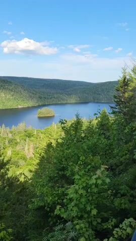 The video shows a landscape with a lake surrounded by green trees. In the middle of the lake, there is a small island covered in trees. The sky is blue with some white clouds. The camera pans slowly from right to left, showing more of the lake and the forested hills in the background. The sun is shining, creating shadows on the trees and water.