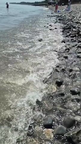 The video shows a pebble beach and the adjacent sea with several people present. The camera is at ground level, providing a close-up view of the sea washing over the pebbles on the beach. The water is clear but slightly murky from the stirred-up sand and pebbles. Several individuals are seen swimming or wading in the water, while others are on the beach, including children. The sky is clear and bright. The scene suggests a typical day at a pebble beach.