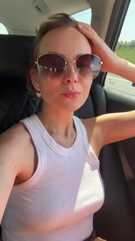 A young woman with blonde hair pulled back, wearing sunglasses and a white tank top, is in the passenger seat of a car. Her right hand is on her head, and her left arm is resting on the armrest. The sun is shining, and the scenery outside the window suggests she is on a highway. She is looking at the camera and smiling.