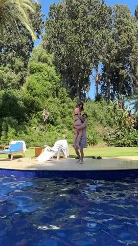 A man holding a woman in his arms jumps into a swimming pool. The man is wearing patterned swim trunks, and the woman is wearing a bikini. They are captured mid-air as they enter the water, creating a splash. The background shows a lush green garden with trees and lounge chairs by the pool. The sun is shining, indicating it is a daytime outdoor scene.
