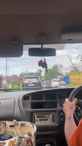 This is a first-person perspective video taken from inside a vehicle, likely a car, looking out through the windshield. The vehicle appears to be stationary or moving slowly in traffic. The dashboard is visible in the foreground, with a stereo system, air vents, and a gear shifter. A tote bag with a cat pattern is also on the passenger side of the dashboard. The driver's hands are visible on the steering wheel. Outside the vehicle, there is a road with other vehicles, including cars and motorcycles. Pedestrians are also present on the side of the road. The scene depicts a busy urban or suburban environment with a cloudy sky. The focus is on the traffic and the surrounding environment as viewed from the car.