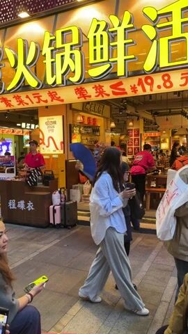 Here's a detailed description of the video:

The video shows a street performance in front of a restaurant. The performer is wearing a traditional Chinese opera costume, complete with a colorful mask (predominantly blue and white), a red robe embroidered with gold, and matching boots. They are holding a blue fan and performing dance-like movements, gesturing to people passing by and seemingly interacting with the staff of the restaurant.

The restaurant has a bright yellow sign with Chinese characters above the entrance. People are seated inside the restaurant. Restaurant staff, wearing red shirts, are attending to customers.

Pedestrians are walking by on the sidewalk. Some people are looking at the performer, and a few are filming with their phones. There are suitcases and bags near the entrance, suggesting some people might be traveling. The scene appears to be taking place outdoors in an urban setting.