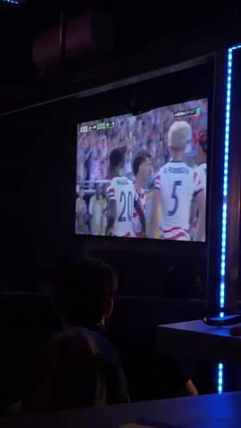 A group of people are in a dimly lit room, watching a large screen that displays a soccer match. The screen shows two male soccer players in red and white striped jerseys with the number 20 visible on one of them. The players appear to be interacting with each other, possibly embracing. There are blue LED lights running vertically along the right side of the screen. In the foreground, partially obscured by darkness, are viewers sitting down, facing the screen. A table is visible in the lower right corner. The overall atmosphere suggests a casual viewing of a sporting event.