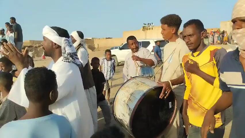 Here is a description of the video:

The video captures a lively street performance in what appears to be a desert-like setting. Several individuals are gathered around a musical ensemble. A man dressed in traditional white attire and a turban is actively singing. Next to him, two other men are engaged in playing percussion instruments, including a large drum. One individual is playing with drumsticks.

The scene is filled with people of all ages, many of whom are looking at the musicians. A car is parked in the background. The overall atmosphere seems celebratory and joyful, as the community appears to be enjoying the performance.