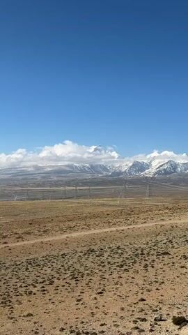 The video captures a wide, expansive view of an arid landscape under a clear blue sky. In the foreground, dry, brown and yellowish scrubland stretches across the frame, marked by a winding dirt track. Several large, metallic electricity pylons stand in the mid-ground, receding into the distance.

Beyond the pylons, a majestic range of snow-capped mountains dominates the background. Their peaks are partially shrouded in white, fluffy clouds. The sun is shining brightly, casting a natural light over the entire scene.

The camera slowly pans horizontally from left to right, revealing more of the vast panorama. Muffled voices are audible in the background, suggesting people are nearby but remain out of frame. The setting indicates a high-altitude, remote region, consistent with Gyirong County, China. The overall impression is one of stark natural beauty and immense scale.