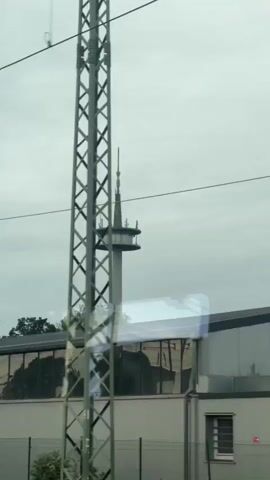 The video shows a view from a moving vehicle, likely a train, looking out a window. In the background, a tall communications tower with a circular platform near the top is visible against a cloudy sky. In the foreground, a long, modern building with a glass facade and a flat roof is seen. Electrical wires and a tall, latticed metal pole are also present in the frame. The reflections on the building's windows obscure parts of the view and create distorted images. The camera pans slightly, suggesting movement.