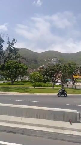 This media file shows a street scene in Catia La Mar, Venezuela. The foreground features a concrete barrier with white lines painted on the road. Beyond the barrier, a multi-lane road is visible with several vehicles. On the left, a person rides a motorcycle. Further down the road, a white car is driving.

In the middle ground, there is a grassy area with trees and a streetlamp. Several houses and buildings are visible on a hillside in the background, suggesting a somewhat dense urban environment against a backdrop of rolling hills. The sky is partly cloudy, indicating it is daytime, likely late morning or early afternoon.

Some construction equipment, including a yellow backhoe, is present on the side of the road, along with orange traffic cones, suggesting ongoing road work or maintenance. A blue truck is also seen driving on the road. The overall atmosphere appears to be a typical day with traffic and some urban activity. No specific emotions are conveyed, and no legible text is visible.