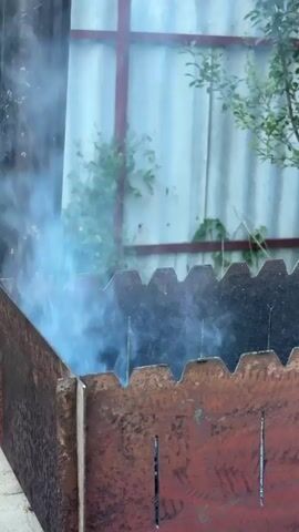 The video shows a close-up of smoke rising from a metal barbecue grill. The grill is made of weathered red metal and has a jagged edge along the top. It is placed on top of a brick structure. In the background, a metal chimney pipe is visible, also emitting smoke. The environment appears to be outdoors, with a fence and some greenery in the background. The lighting suggests it is daytime.