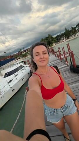 This is a short video clip featuring a woman taking a selfie on a wooden pier. She is wearing a red bikini top and denim shorts, and she is smiling at the camera while raising her right arm. The pier extends into a body of water, likely the sea, and a white catamaran is docked to the left. In the background, there are palm trees, buildings, and a cloudy sky. The overall impression is a vacation or travel scene, likely during the day given the light, though the clouds suggest it might be overcast or nearing sunset. The woman appears happy and relaxed, enjoying her surroundings. There is no visible text in the video. The location is identified as Baan Bang Rak, Thailand.