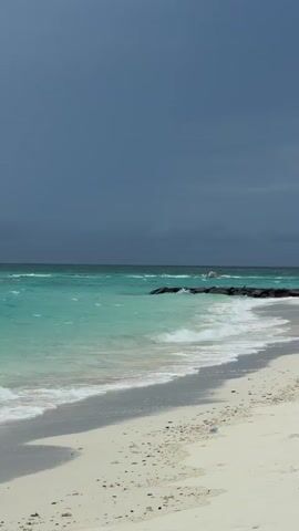 The video shows a beach with white sand and light turquoise water. Dark, ominous clouds fill the sky, hinting at an approaching storm. A rocky barrier extends slightly into the ocean, breaking the waves. The waves are moderate and create white foam as they crash onto the shore. No people or other moving objects are visible.
