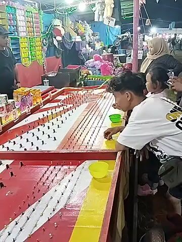 A man plays a carnival game that involves dropping coins into slots. He’s standing at a tall, horizontal machine with rows of pegs and red, white, and yellow stripes along the bottom. He holds a small green bowl containing coins. There’s a row of prizes behind the game machine, and other people are in the background. The scene takes place at night with some visible lighting and neon decorations.