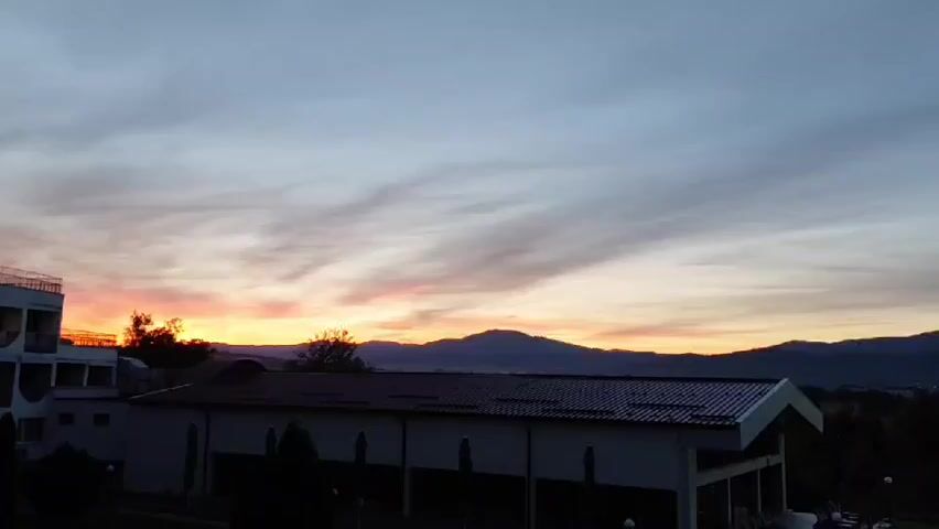 This video captures a time-lapse of a sunrise over a mountainous landscape. In the foreground, there are several buildings, including a white multi-story structure on the left and a long, rectangular building with a dark roof in the center. The central building appears to be a facility with outdoor seating and lounge chairs arranged around a pool area. The sky transitions from a pale blue and grey with wisps of clouds to vibrant oranges and pinks as the sun rises, casting a warm glow over the mountains in the distance. Trees and greenery are visible around the buildings, and the overall atmosphere suggests an outdoor resort or a similar recreational area. There are no visible people or animals in the footage.