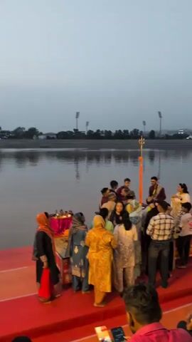 A crowd of people are gathered on a red carpeted area next to a body of water. Many of the people are wearing traditional Indian clothing. Some individuals are standing near small tables with what appear to be religious items. In the background, there is a body of water, a shoreline with trees and structures, and a hazy sky. There are lights visible along the shoreline in the distance. The overall atmosphere suggests a cultural or religious gathering.