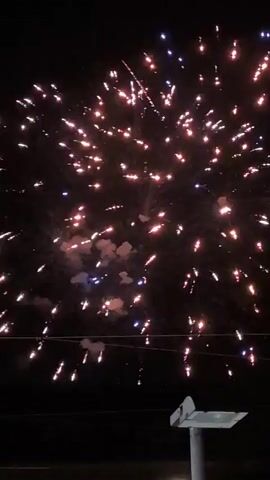 The video captures a fireworks display at night. The sky is dark and filled with numerous bright explosions of light and smoke. Fireworks ignite and burst in various patterns and colors, predominantly oranges and whites, with some blue sparks visible. A single streak of light, possibly from a firework rocket, ascends in the upper left portion of the frame. Below the fireworks, dark silhouettes of trees and power lines are visible against the illuminated sky. In the foreground, a portion of a white object, possibly a light fixture or street sign, is visible on the right side of the frame. The overall scene depicts a festive and energetic fireworks celebration.