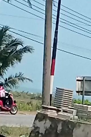 A red car is visible passing by a utility pole. The pole has red, white, and black markings. There are power lines and palm trees in the background, suggesting an outdoor setting. There is a stack of flat objects near the base of the pole, and some construction materials are also visible.