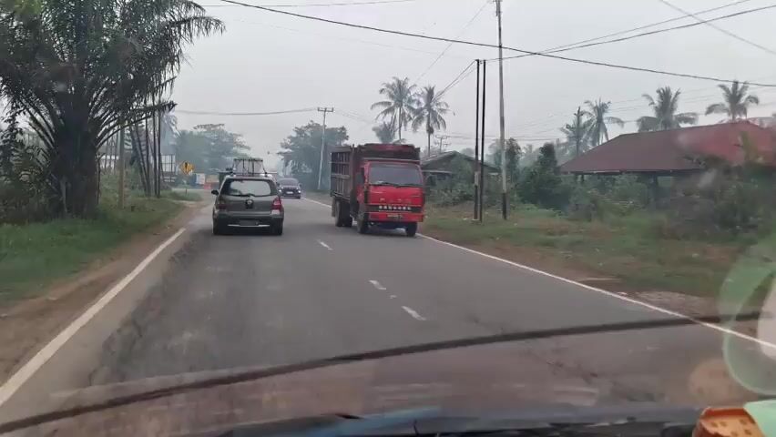 The video shows a road scene in what appears to be a rural or semi-rural area. The sky is overcast, and there is some haze or smoke in the air, suggesting potential air pollution or natural conditions like fog or dust.

On the road, there are several vehicles. In the foreground, we see the dashboard and windshield of a vehicle from which the video is presumably being recorded. To the left of the road, there are palm trees and other vegetation.

The main focus of the video is the traffic on the road. Several cars are visible, including a dark colored hatchback in the left lane, a black SUV in the right lane, and a white SUV further ahead in the right lane. There is also a large red dump truck traveling in the right lane, ahead of the dark hatchback and to the left of the black SUV. A motorcycle with a rider is visible on the right side of the road, moving in the same direction as the cars.

Along the sides of the road, there are utility poles with numerous wires, and some buildings or structures, including what looks like a house with a red roof on the right side. There are also some signs visible on the left side of the road. The road itself is paved and appears to be two lanes wide, with a white line marking the center.

The overall impression is of a moderately busy road in an area with tropical vegetation, under a hazy sky. The video is in motion, indicating that the recording vehicle is moving along the road.