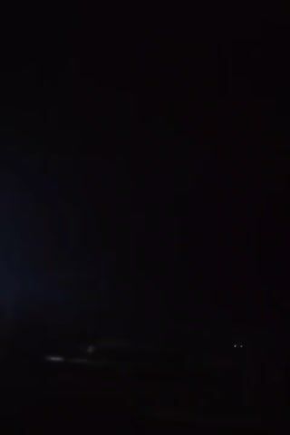 The video captures a dark, nighttime scene with minimal light. A vehicle's headlights are visible, illuminating a section of a road or open area. Distant lights, possibly from buildings or other vehicles, are faintly discernible. The overall ambiance is one of darkness and limited visibility, characteristic of a nighttime environment.