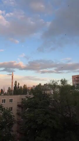 A video shows a view of an urban landscape at sunset. The sky is a mix of blue and gray clouds, with streaks of pink and orange light indicating the setting sun. In the distance, a tall telecommunications tower stands out against the horizon. Below the sky, a cluster of trees and buildings can be seen. The buildings appear to be apartment blocks, some with visible balconies. The overall impression is a quiet evening scene in a city.