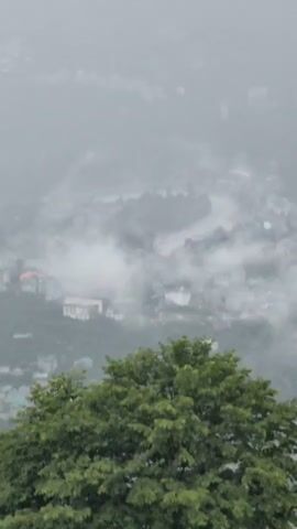 The video captures a misty mountainous landscape with a town nestled in the valley below. In the foreground, a large, lush green tree dominates the lower portion of the frame. The middle ground is obscured by dense fog and clouds, which partially hide the buildings and streets of the town. Some structures, possibly residential or commercial buildings, can be seen scattered throughout the valley, some with distinctive green roofs. A winding river or road appears to snake through the town, also partially veiled by the mist. The overall atmosphere is serene and ethereal, with the fog creating a sense of depth and mystery. The colors are muted, with greens of the foliage contrasting with the grays and whites of the mist and the muted tones of the town. The camera appears to be positioned at a high vantage point, looking down upon the scene.