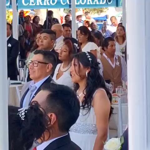 The video shows a wedding celebration. People are dressed formally and seated at tables. The sign "DE CERRO COLORADO" is visible in the background. A woman in a white dress with a sparkling belt and a floral headpiece is standing and smiling. A man with glasses in a suit is sitting next to her. The overall atmosphere seems cheerful and celebratory. The video is filmed through white posts, providing a framed view of the scene.