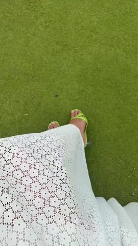 The video shows a person from the knees down, wearing a white lace dress and bright green heeled sandals. The person is walking on a grassy surface, likely artificial turf. The camera angle is from above, looking down at the person's legs and feet. The person's right foot is in the foreground, with the left foot slightly behind and to the left. The lighting is bright, suggesting it is daytime. The background is a blurred green expanse.