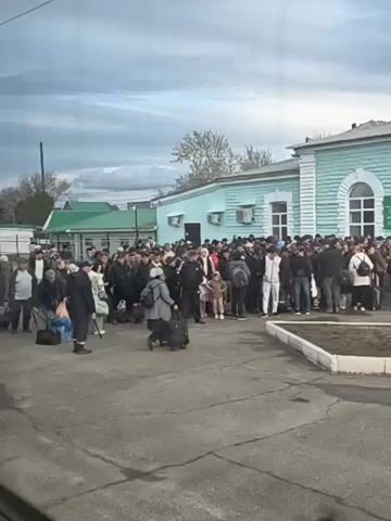 A crowd of people, mostly dressed in dark or neutral-colored clothing, stand outside a light blue building with white trim and windows. Many are holding or pulling luggage.  The people are standing on an asphalt surface. In the background, there's a hint of greenery and a cloudy sky. The video is taken from inside a vehicle looking out.
