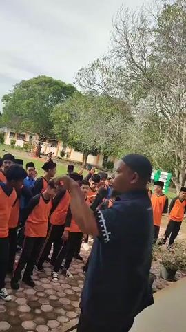 A group of young males, likely students, wearing orange and black uniforms are gathered outdoors. They are standing in a semi-circle, facing a man who is holding a microphone and speaking. The man is wearing a dark blue shirt and a cap. Another person, wearing a headscarf, is partially visible on the left side of the frame. The environment appears to be a schoolyard, with a building in the background and trees. The ground is paved with interlocking bricks. The sky is overcast.