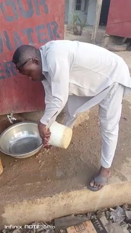 A man wearing a light gray outfit and sandals is outside, bending over a metal bowl to pour water from a white container. He appears to be filling the bowl from a tap or source on a red and blue structure with some writing on it. The ground is wet and dirty around the bowl. There's trash on the ground nearby. A building is visible in the background.