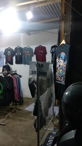 The video captures an indoor or covered outdoor retail space, likely a stall or small shop, in Kecamatan Ciawi, Indonesia. The scene is dominated by a display of numerous t-shirts of various colors and designs. Some t-shirts are hung individually on a light-colored wall, while others are neatly folded and stacked on multiple metal clothing racks.

A bright artificial light source, possibly a bare bulb, illuminates the space from the left side, casting some glare. The ceiling appears to be made of corrugated metal, and the floor is concrete.

Several price tags are visible, written in Indonesian Rupiah (IDR). One tag prominently displays "AS 35 000" near a section of hanging t-shirts. Another tag reads "TAS" (Indonesian for bag) with a price of "160 000," indicating bags are also for sale, possibly on a shelf behind the t-shirts. Further along, a tag on a clothing rack shows "RP 160 000" and "3 PCS," suggesting a multi-pack deal for certain items. A partially obscured banner or sign with text and numbers like "085..." and "000" is visible on the far left.

No people are visible in the frame, and there is no discernible activity other than the presentation of merchandise. The artificial lighting suggests it might be evening or night.