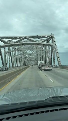 The video captures a first-person perspective of driving across a large metal truss bridge on a cloudy day. The bridge has multiple lanes of traffic, and the camera vehicle is in the right lane. Ahead, several cars and a semi-truck are visible. The structural elements of the bridge, including the steel beams and rivets, are prominently featured. The video is shot from inside a vehicle, with the dashboard and windshield visible in the foreground, reflecting the gray sky and bridge structure. The overall mood is neutral, depicting a typical travel scene on a highway bridge.
