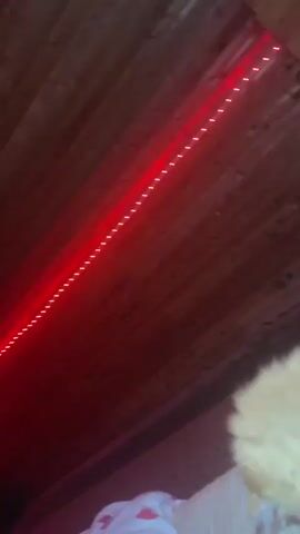 A close-up, slightly blurred, low-angle video captures an orange cat with its head down, interacting with a white earbud cord. The cat is on a bed with a white and pink heart-patterned pillow. The background shows a wooden ceiling with red lighting. The cat's fur is fluffy and the earbud cord is draped over the bed. The cat appears to be chewing or playing with the cord.